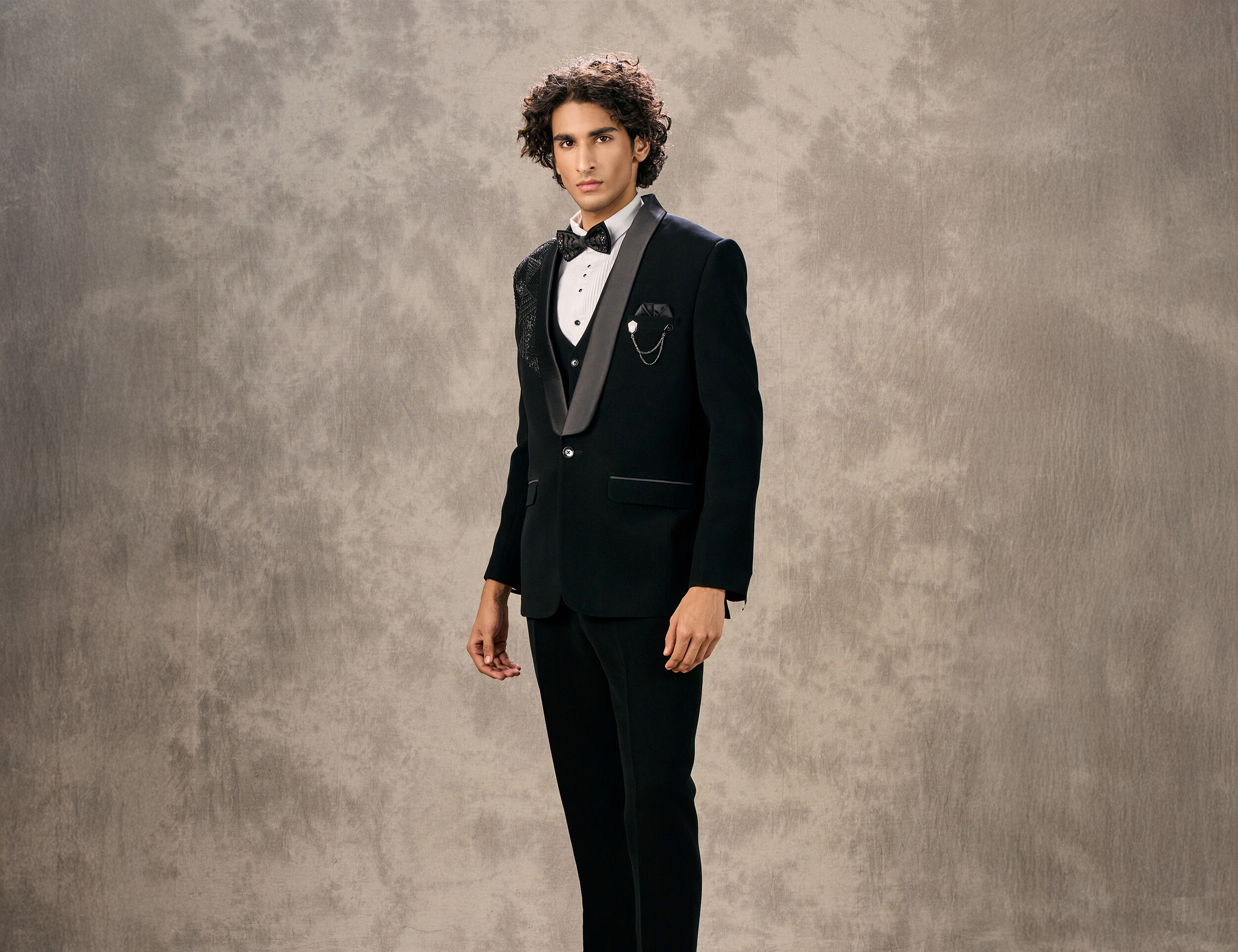 Man in a black tuxedo with satin lapels, a white waistcoat, and a bow tie, showcasing formal wear.
